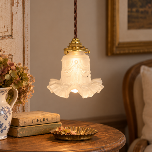 A vintage-style pendant light with a ruffled white glass shade and brass hardware, hanging over a wooden table in a cosy, decorated room.