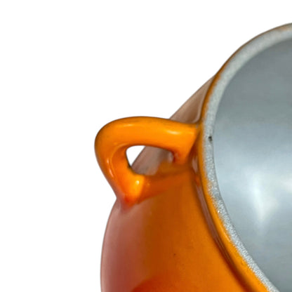 Close-up of a glossy orange ceramic pot or casserole dish showing the side handle, interior rim, and light gray interior surface against a white background.