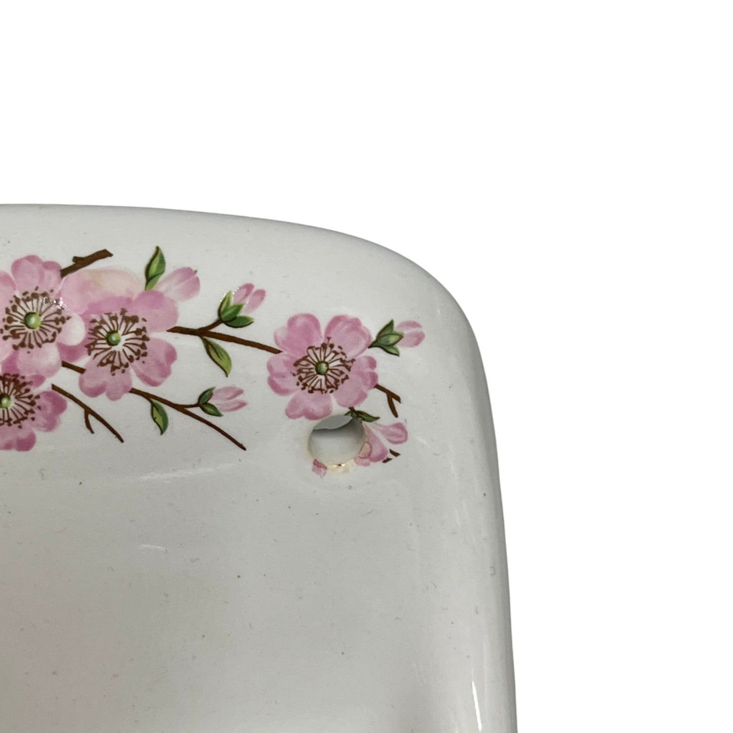 Close-up of a white ceramic porcelain sink basin corner decorated with pink cherry blossom flowers on a branch, featuring a drain hole, against a white background.