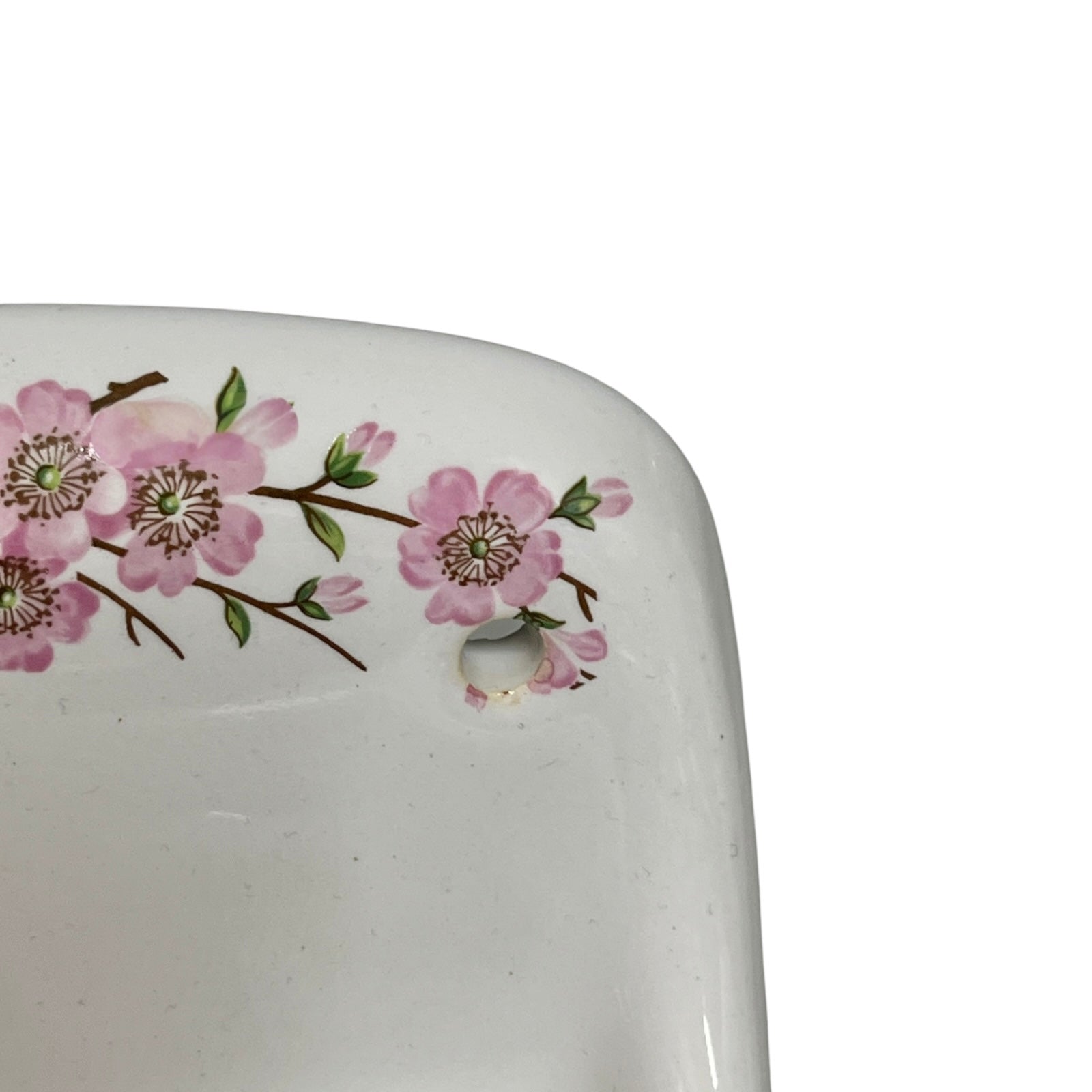 Close-up of a white ceramic porcelain sink basin corner decorated with pink cherry blossom flowers on a branch, featuring a drain hole, against a white background.