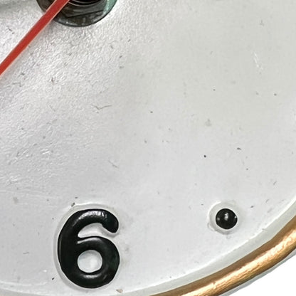Close-up of a white clock face showing the number 6 in black, with a gold-tone rim edge and part of a red second hand visible.