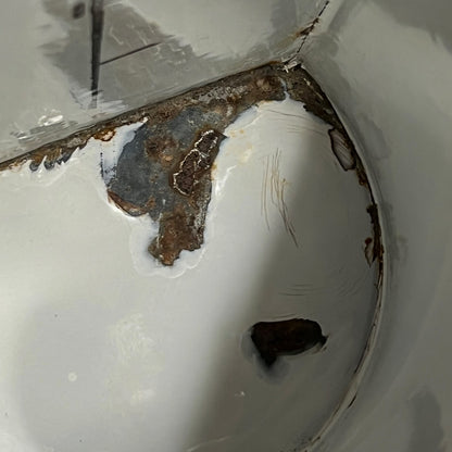Close-up of a white enamel pot showing heavy rust and corrosion around the rim and inside the bowl, with a visible drain hole.