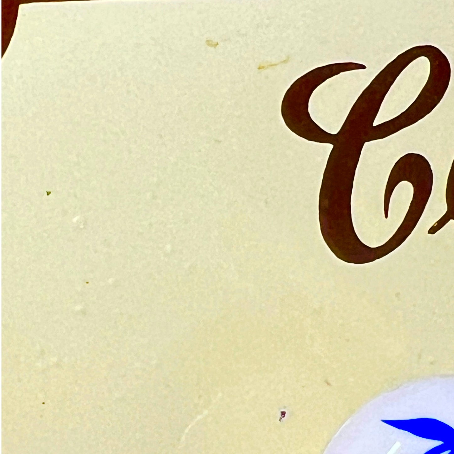 Close-up of a cream-colored surface with partial dark brown cursive lettering and a small blue-and-white circular object at the bottom right.