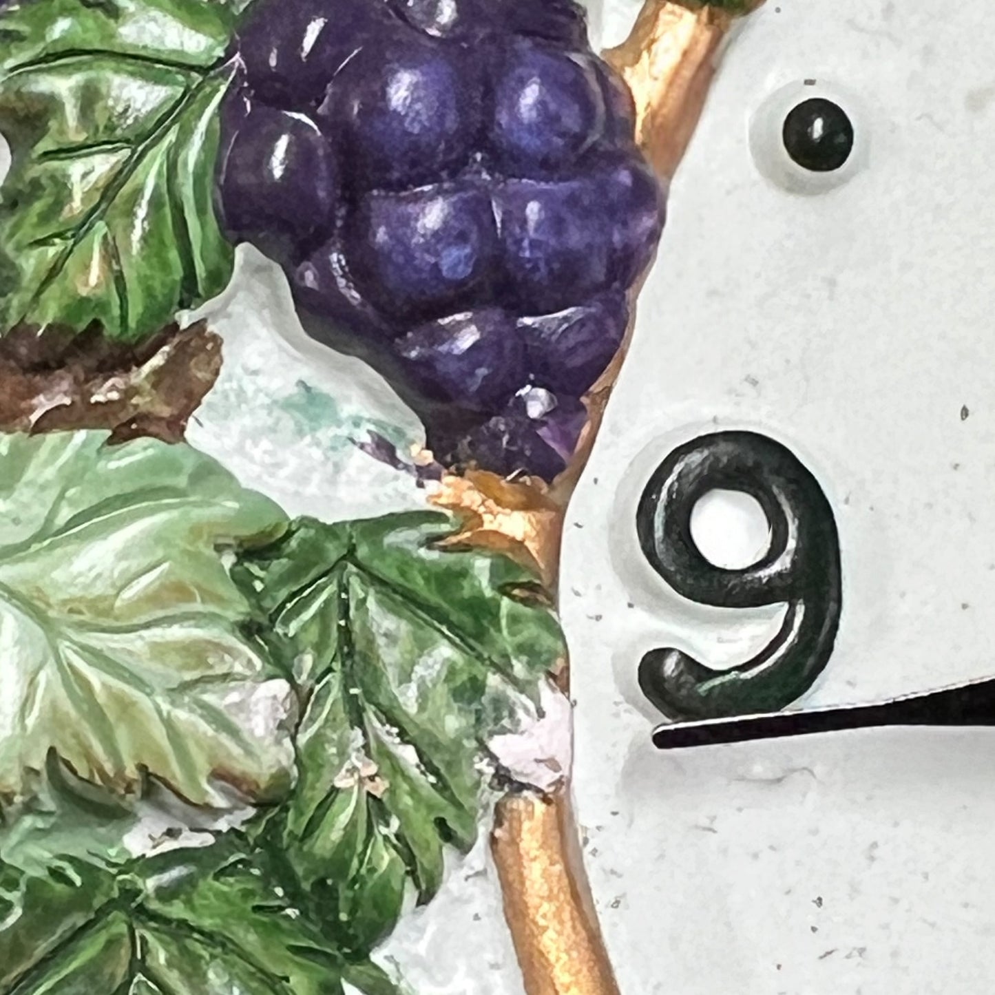 Close-up detail of a decorative wall clock face featuring a raised purple grape cluster with green leaves and a gold vine, alongside a black numeral 9 on a white textured background.