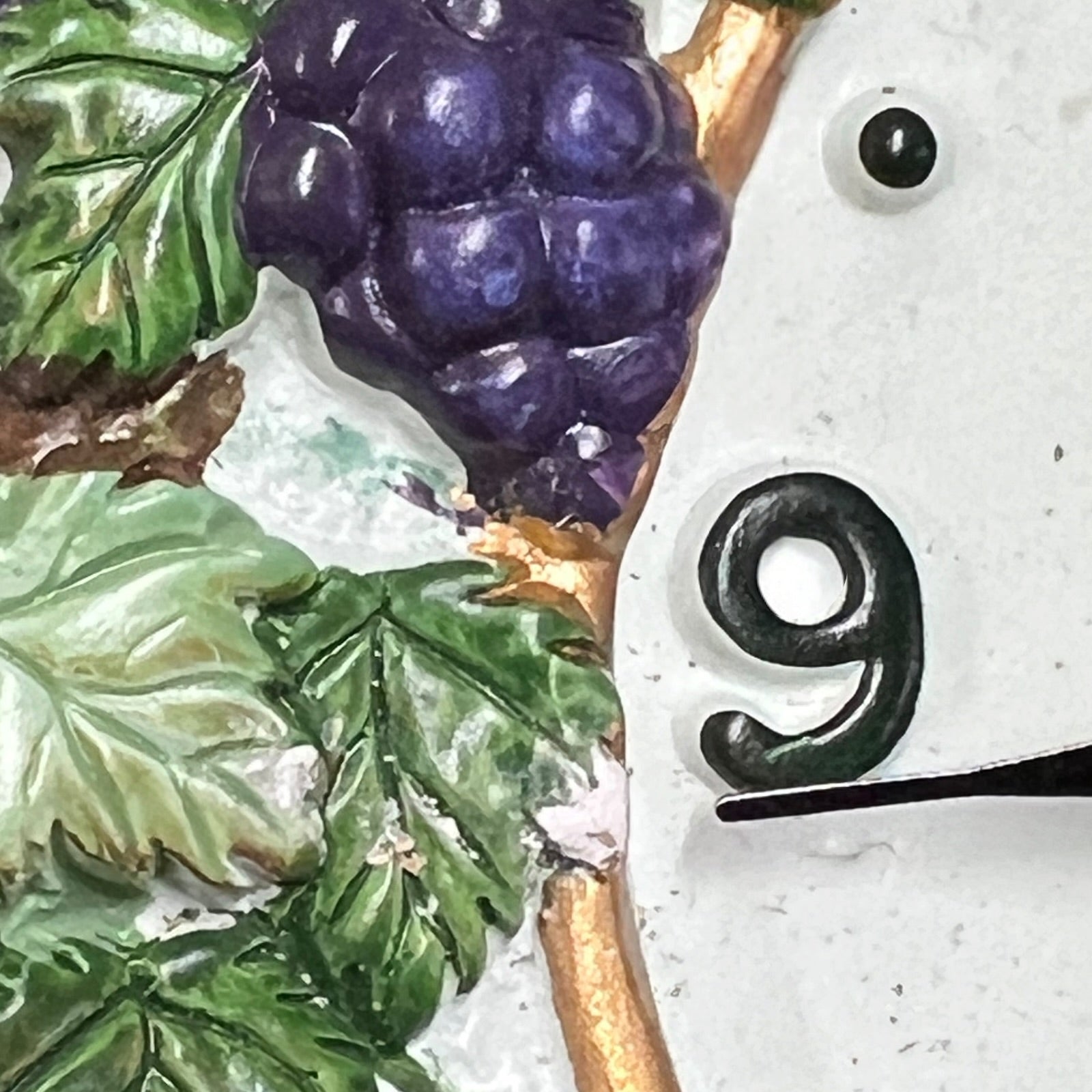 Close-up detail of a decorative wall clock face featuring a raised purple grape cluster with green leaves and a gold vine, alongside a black numeral 9 on a white textured background.