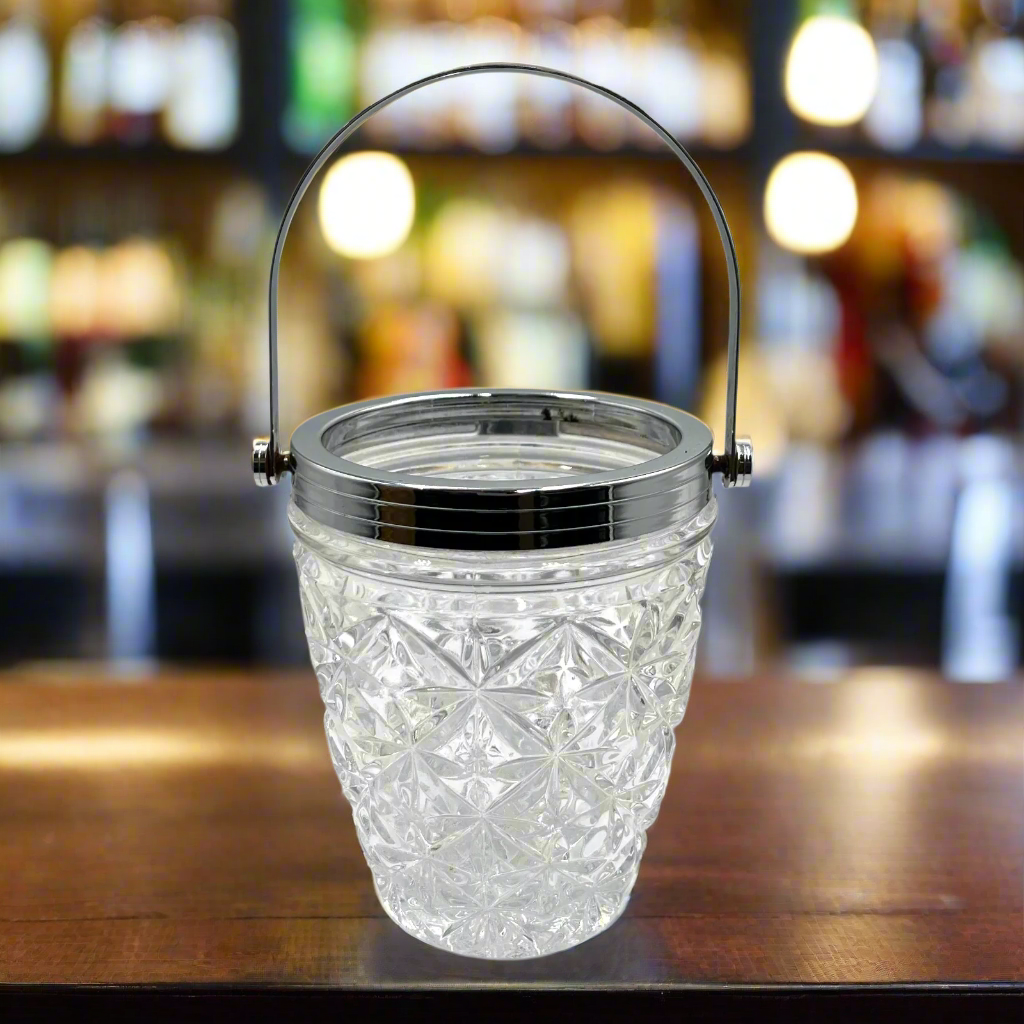 Decorative glass ice bucket with handle on a wooden surface, blurred bar background