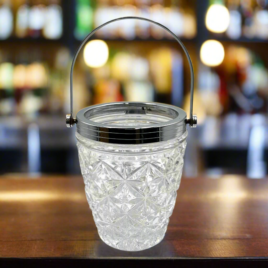 Decorative glass ice bucket with handle on a wooden surface, blurred bar background