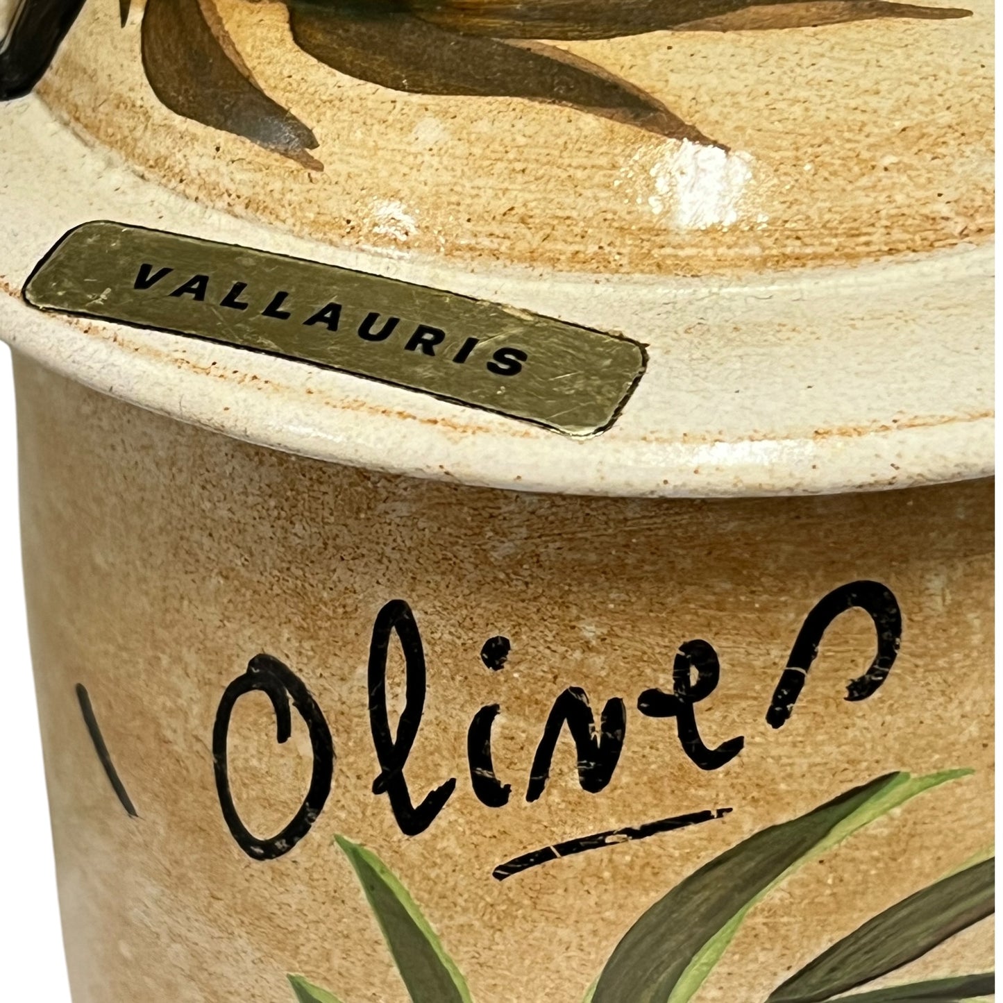 Close-up of a ceramic olive jar with a matching lid, featuring the brand name 'VALLAURIS' on a metal plaque and 'Olive' written in black script on the side, decorated with olive leaves.