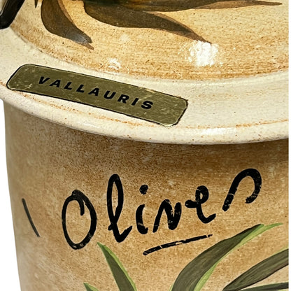 Close-up of a ceramic olive jar with a matching lid, featuring the brand name 'VALLAURIS' on a metal plaque and 'Olive' written in black script on the side, decorated with olive leaves.