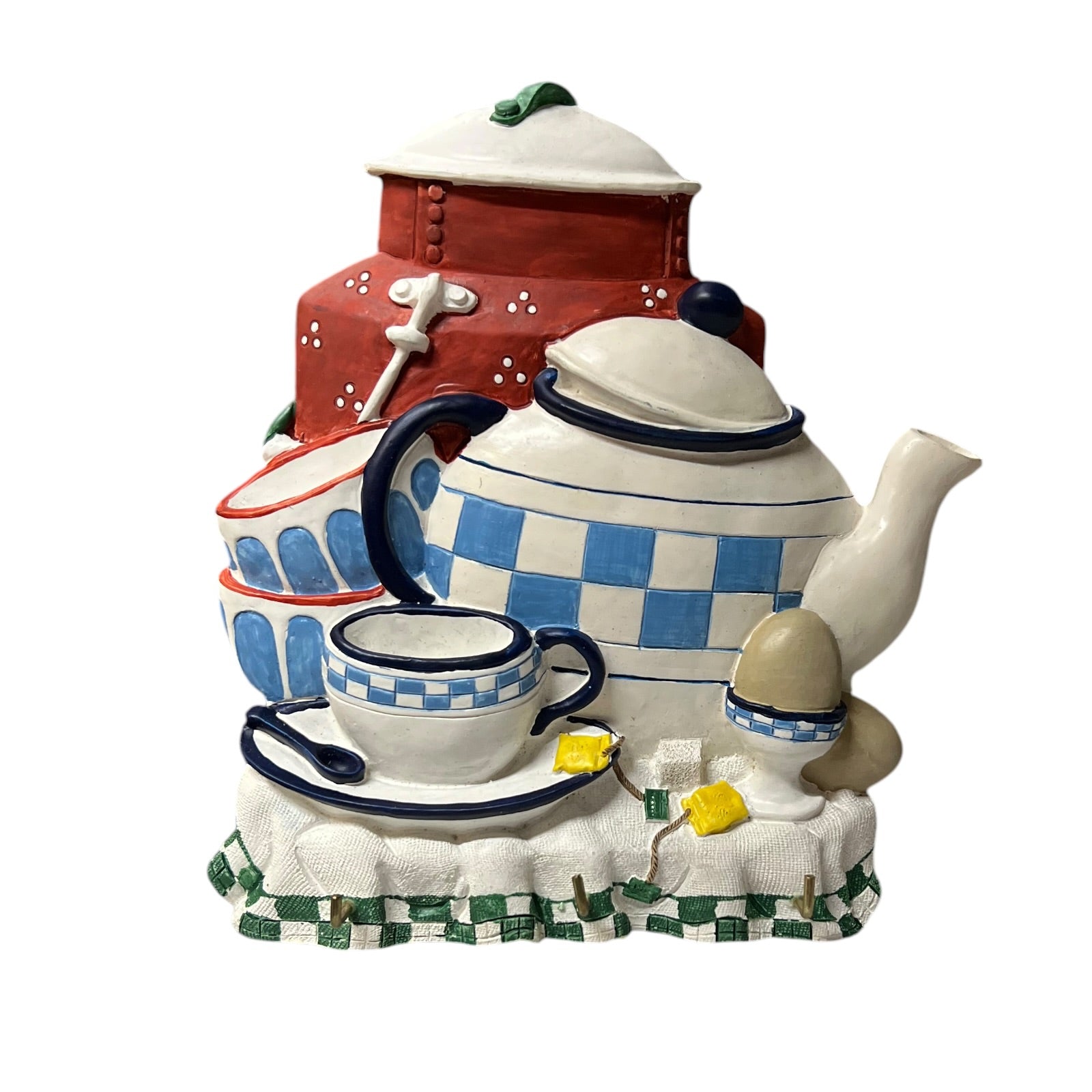 A ceramic kitchen-themed decorative sculpture featuring a white teapot with blue checkers, a white teacup and saucer with blue trim, a red storage chest, and a white egg on a patterned base against a plain white background.