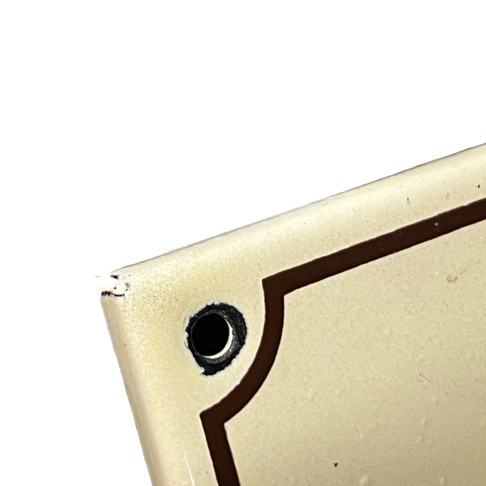 Close-up of the corner of a cream-colored metal panel with a pre-drilled hole and a dark brown decorative border, set against a plain white background.