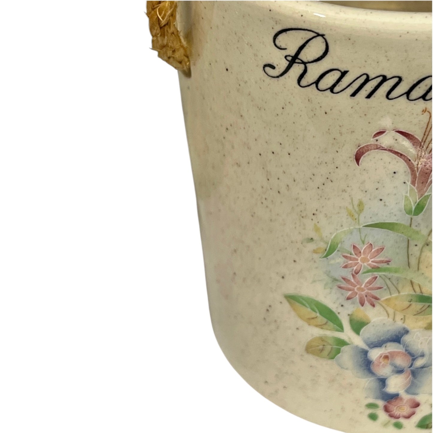 Close-up of a speckled cream ceramic container with a gold-toned handle and floral decoration, including the partial script text 'Rama.