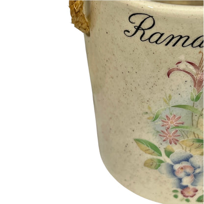 Close-up of a speckled cream ceramic container with a gold-toned handle and floral decoration, including the partial script text 'Rama.