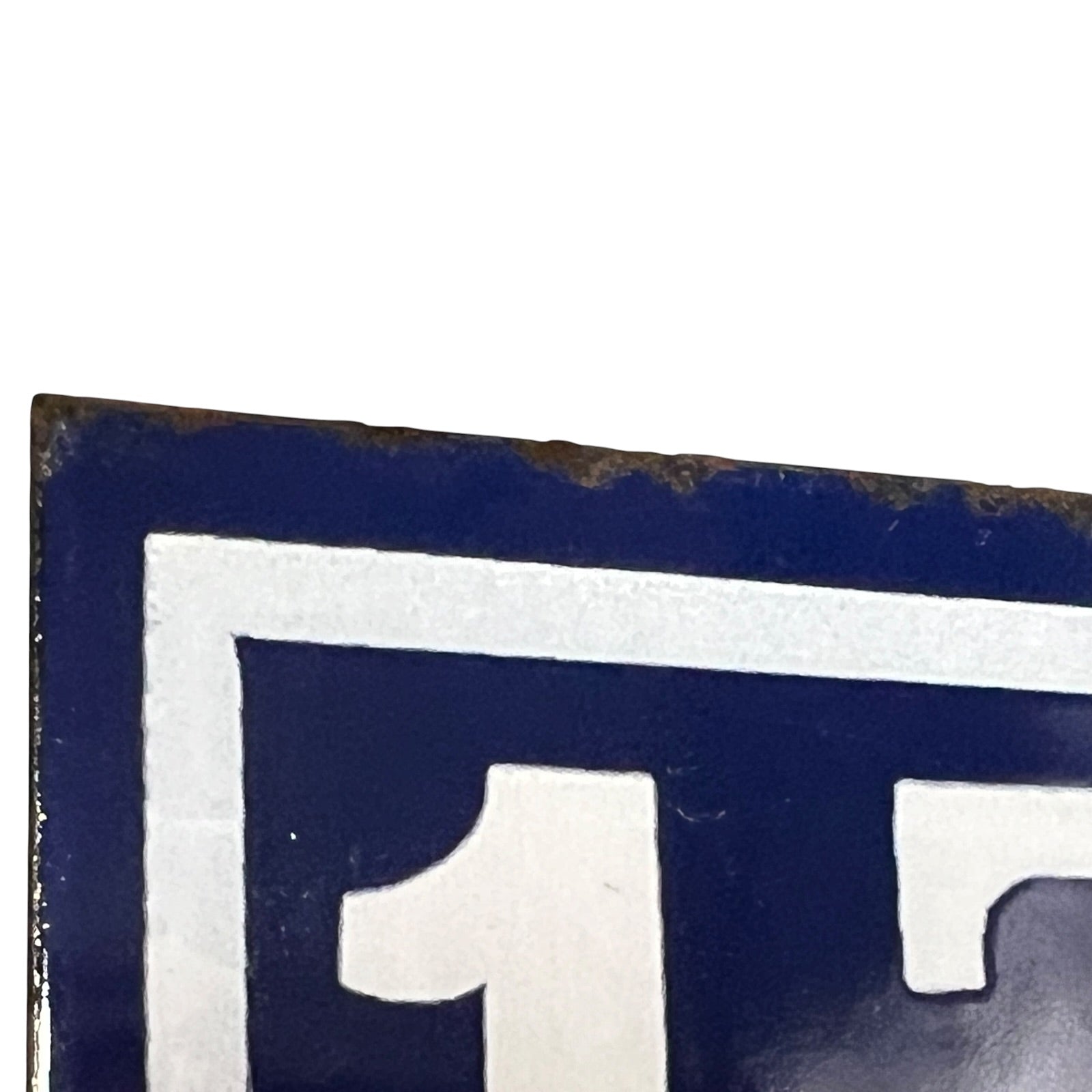 Close-up of a dark blue and white metal sign showing part of a bold white numeral on a dark blue background, with some rust along the top edge.