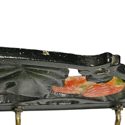 Close-up of a black cast iron decorative tray with hand-painted fish motifs in orange, pink, and green, supported by brass legs against a white background.