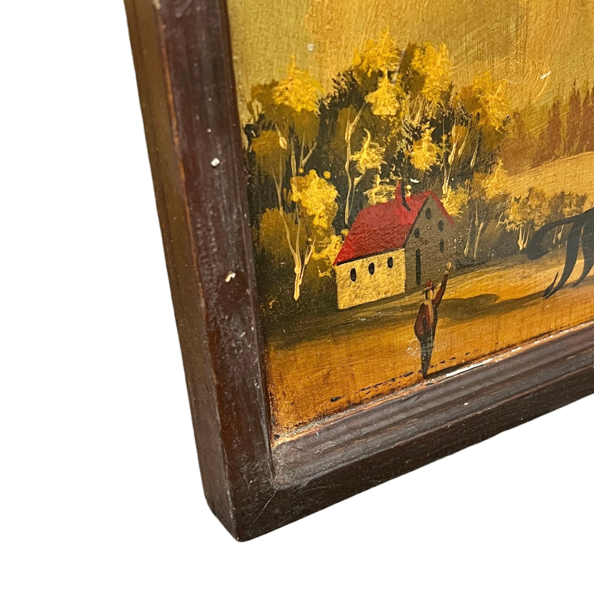 Close-up corner view of a framed rustic landscape painting showing a small house with a red roof, yellow trees, and a dark brown wooden frame on a white background.