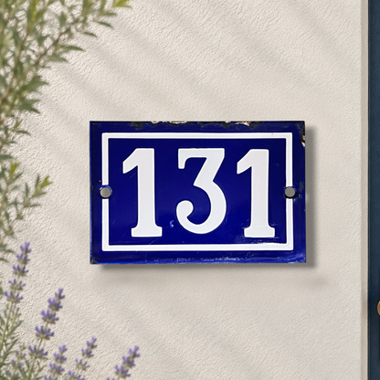 A rectangular blue house number plaque with white numerals '131' mounted on a textured off-white wall, with some greenery and lavender visible in the foreground.