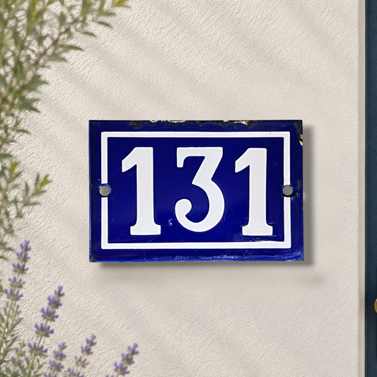 A rectangular blue house number plaque with white numerals '131' mounted on a textured off-white wall, with some greenery and lavender visible in the foreground.