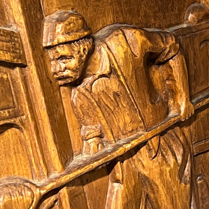 Wooden carving of a person with a hat and long coat holding a staff.