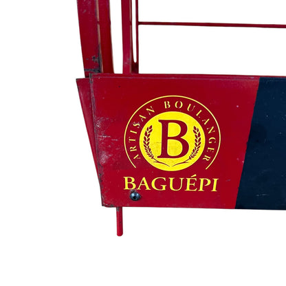 A close-up of a red metal bakery display stand or counter panel with a yellow circular logo reading 'Artisan Boulanger B Baguépi'.