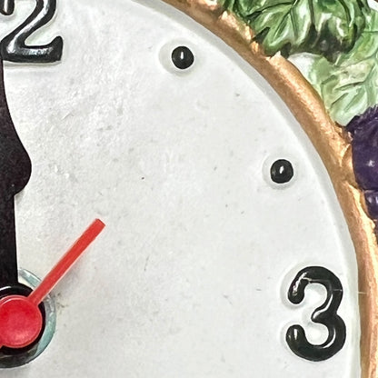 Close-up detail of a decorative clock face with a white dial, black numerals, black dot hour markers, a red second hand, and a gold-toned border with green and purple painted accents.