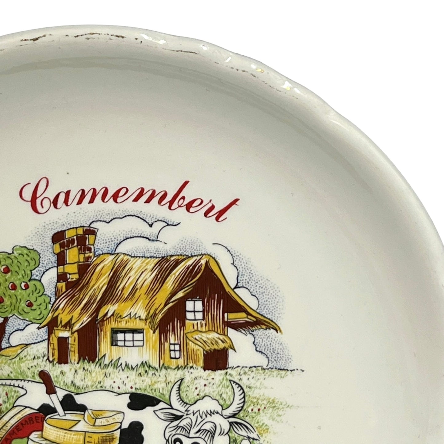Ceramic plate with a illustration of a thatched roof house and cow, and the word 'Camembert' on a white background.