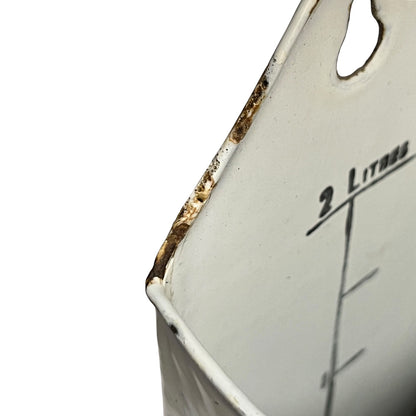 Close-up of a white enamel measuring container with a 2-liter mark and black measurement lines, showing worn rusted edges against a white background.