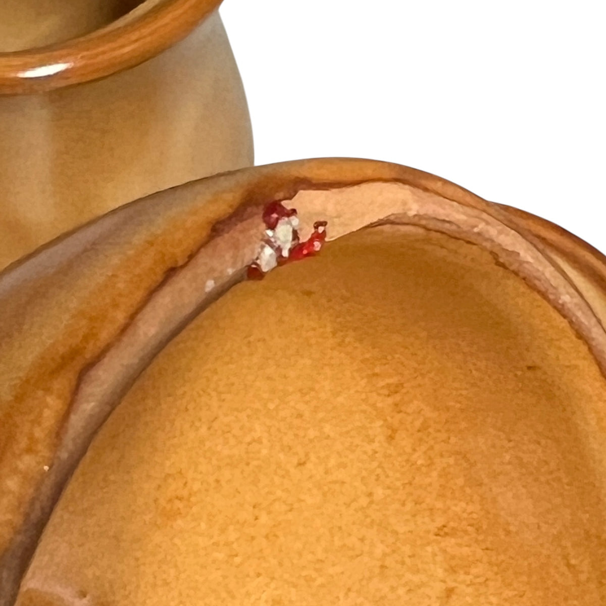 Close-up of a ceramic jar with a warm orange-brown glazed finish, showing the rim and a small red and white decorative detail, against a white background.
