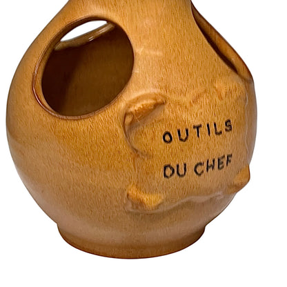  side view of a vintage French ceramic kitchen utensil holder with a honey-brown mottled glaze, rounded body and two large angled side openings. Raised front plaque inscribed Outils du Chef, meaning Chef's Tools. Traditional rustic French country kitchen pottery measuring 18.5 cm high, sourced from a French brocante and shown on a plain white background 