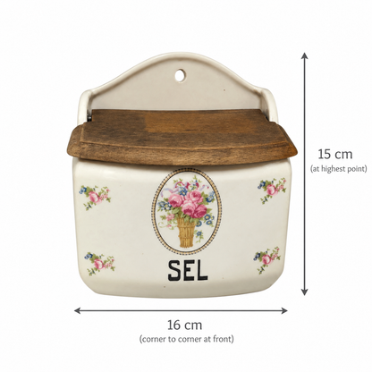 An off-white ceramic wall-mounted storage box with a dark brown wooden lid, decorated with a floral bouquet design and the letter 'SEL'. Black dimension lines and text indicate its height is 15 cm and width is 16 cm.