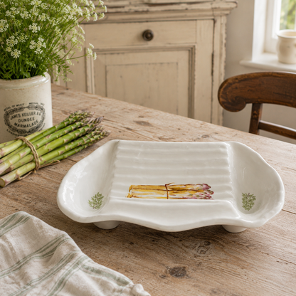 A white ceramic serving platter with a ribbed interior and asparagus illustrations near the corners, displayed on a rustic wooden table with asparagus and a potted plant in the background.