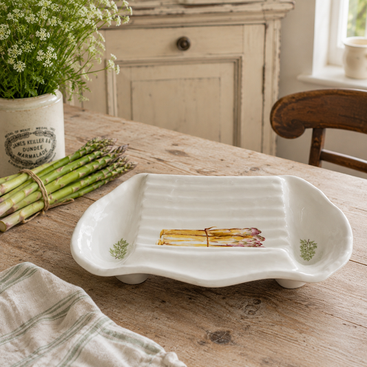 A white ceramic serving platter with a ribbed interior and asparagus illustrations near the corners, displayed on a rustic wooden table with asparagus and a potted plant in the background.