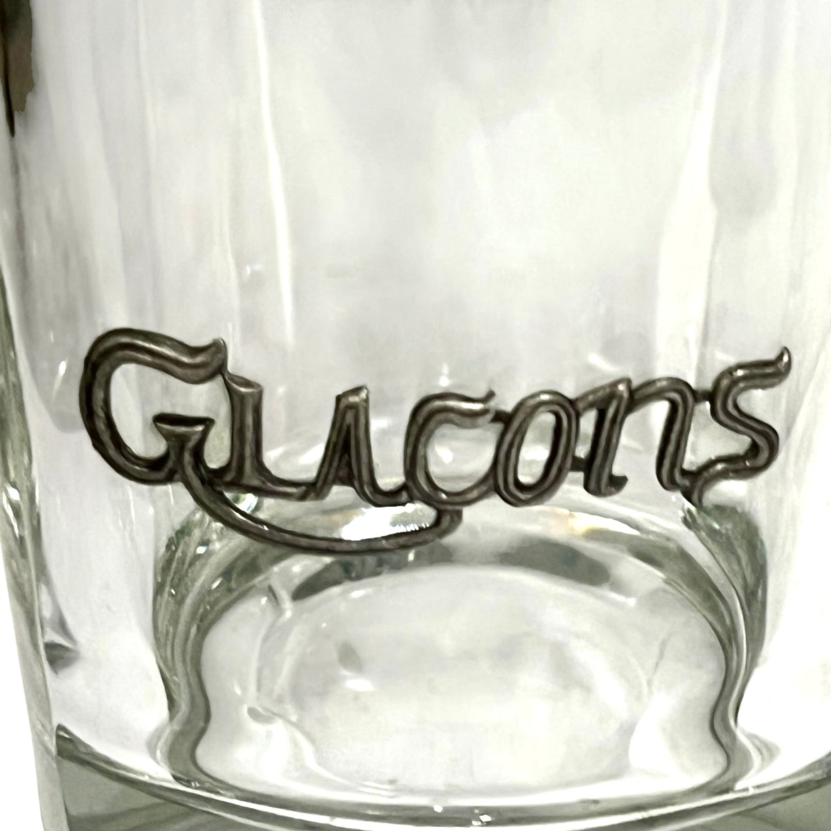 A close-up of a clear glass ice bucket featuring a raised metallic script logo that reads 'Glacons' in a dark silver or gunmetal finish.
