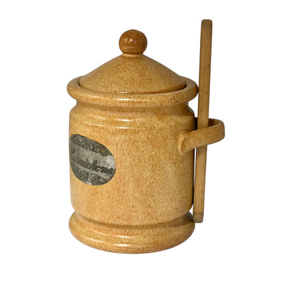 A small beige ceramic lidded jar with a wooden tongue pair resting in its side handle, featuring a round wooden knob on top and a decorative oval label on the front.