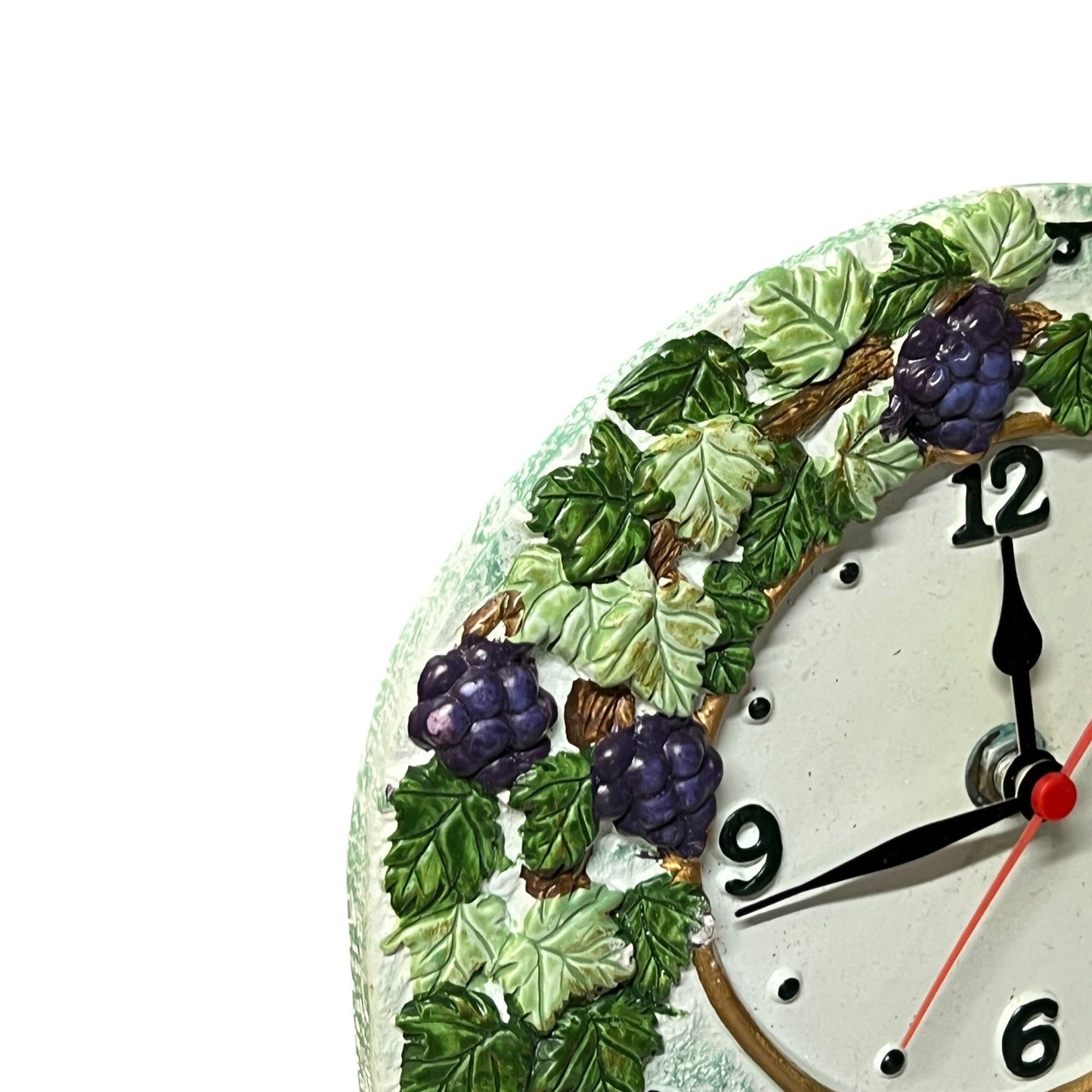 Close-up of a decorative wall clock face showing the numbers 12, 9, and 6, with black hands and a red second hand. The clock is decorated with raised green leaves and purple grapes around the edge, on a white background.