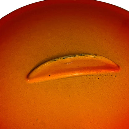 A close-up of a glossy orange-red bean pot, possibly a small accessory or component, set against a matching orange background.