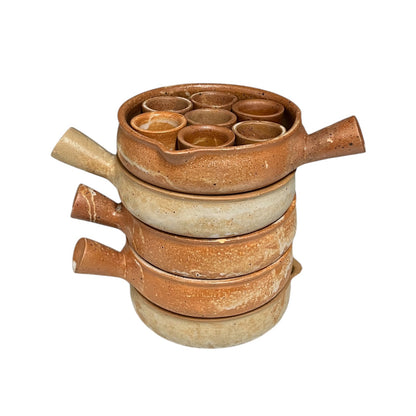 A handmade terracotta clay set of stacked shallow bowls and cups with small side handles, featuring a top tray with six small cup compartments, photographed on a solid white background.