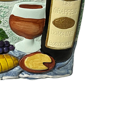 A close-up of a decorative ceramic wine-themed plaque or tray featuring a wine bottle labeled Les Grappes Domaine Bordeaux, a filled wine glass, grapes, cheese, and a yellow bowl on a blue-gray textured base against a white background.