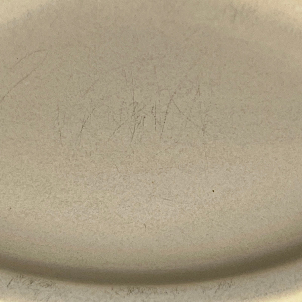 Close-up of a light beige ceramic surface with faint pencil-like markings and a curved rim visible at the bottom edge.