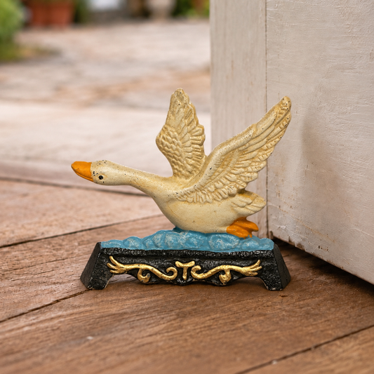A decorative cast-iron duck door stop with wings raised, standing on a blue and gold wave-like base. The duck has a cream body with orange beak and feet, photographed outdoors on a wooden surface next to a light-colored wall.