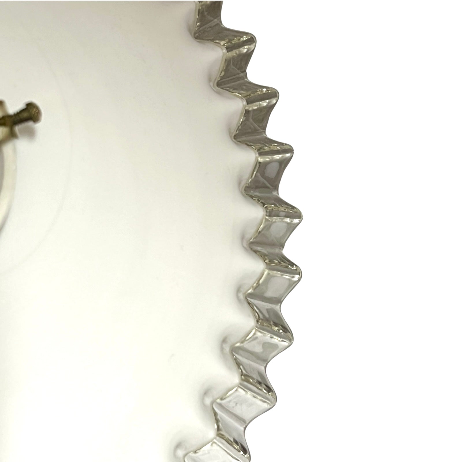 A close-up of a clear, wavy-edged glass container or bowl on a plain white background, showing the reflective silver-toned rim and a small portion of a metallic component at the left edge.