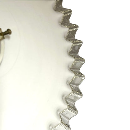 A close-up of a clear, wavy-edged glass container or bowl on a plain white background, showing the reflective silver-toned rim and a small portion of a metallic component at the left edge.