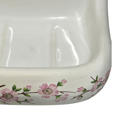Close-up of a white ceramic dish or bowl with a pink floral branch pattern around the rim, shown against a white background.