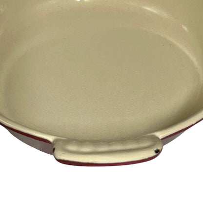 Close-up of a round baking dish or casserole dish with a cream-colored enamel interior and a dark red rim, set against a plain white background.