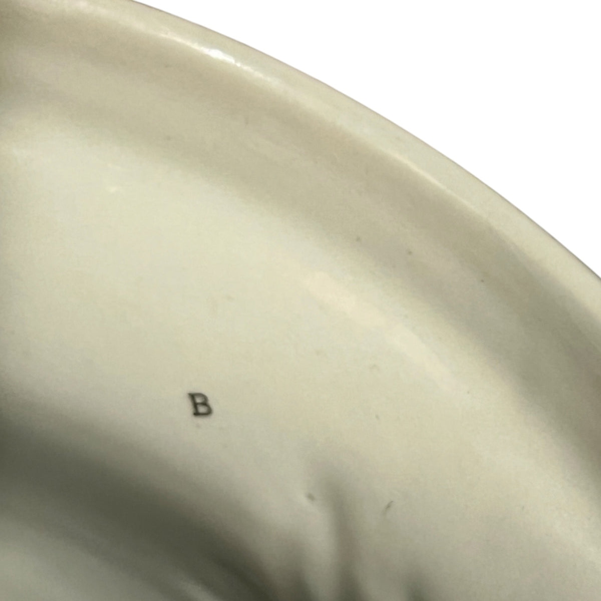Close-up of the interior of a glossy off-white ceramic plate or dish, showing a small dark letter 'B' mark on the surface against a white background.