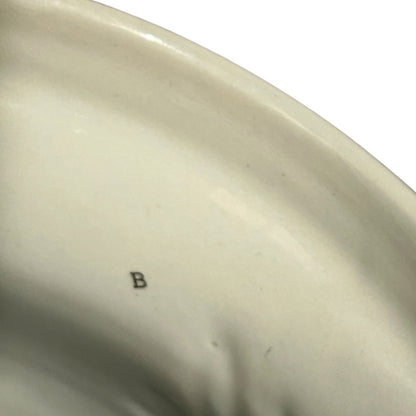 Close-up of the interior of a glossy off-white ceramic plate or dish, showing a small dark letter 'B' mark on the surface against a white background.