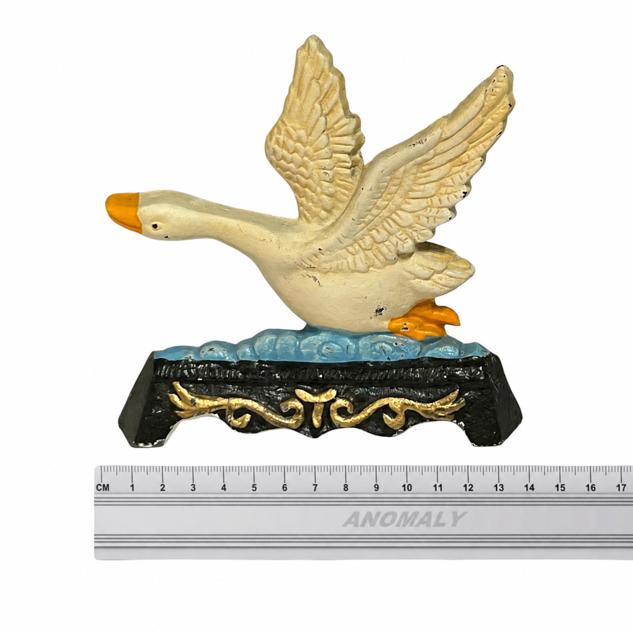 A decorative goose figurine with outstretched wings on an ornate black and gold base, shown above a metal ruler for scale on a white background.