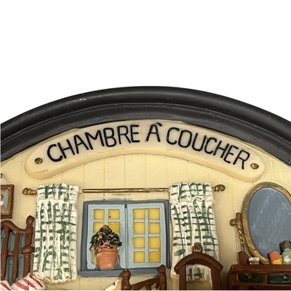 A close-up of a miniature decorative figurine representing a child's bedroom, featuring the French text 'CHAMBRE A COUCHER' on a cream-colored arched wall above a window with blue shutters and green-and-white patterned curtains.