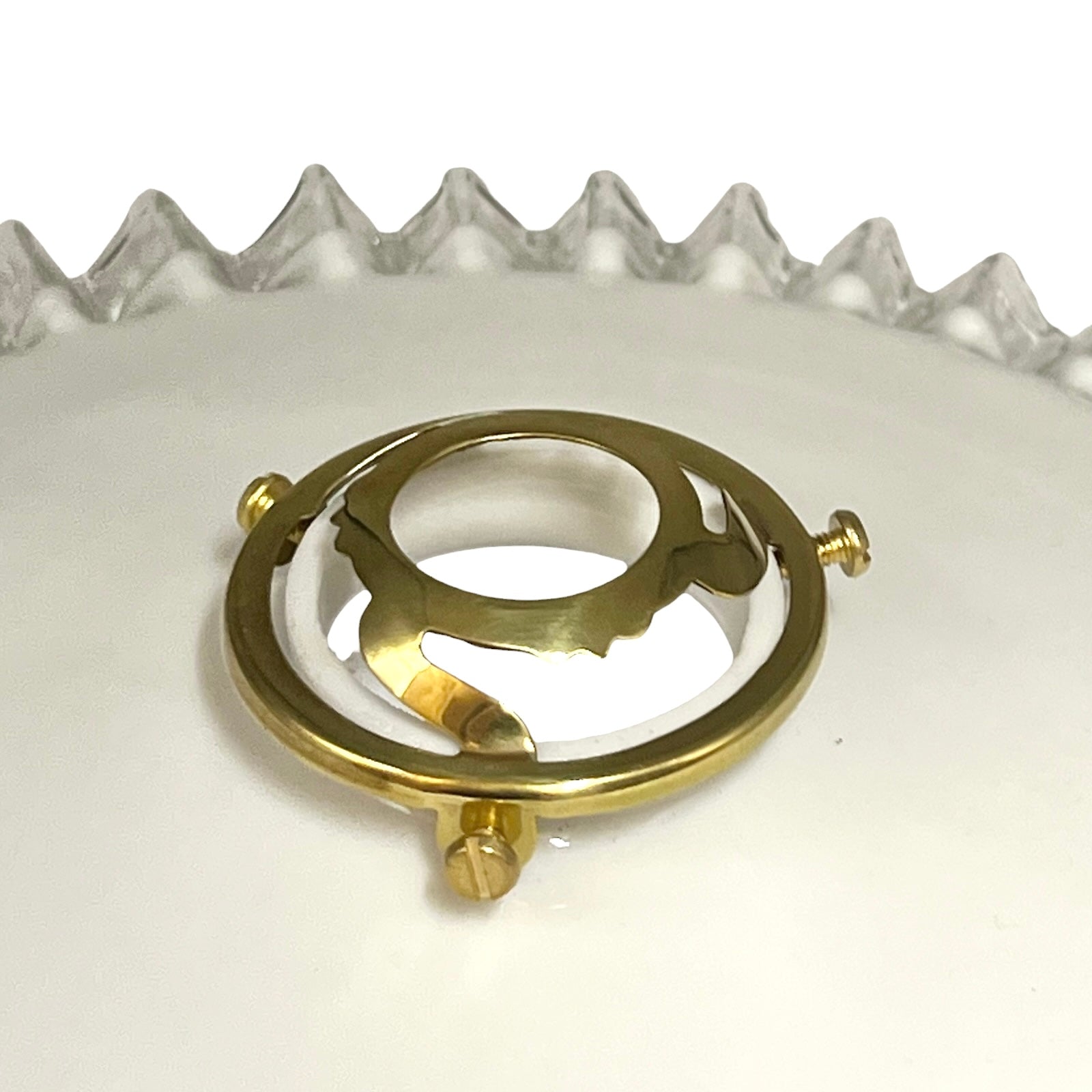 A polished gold metal ring stand with a circular design and three screw posts, displayed on a white surface with a glass lampshade in the background.