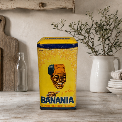 A vintage-style rectangular tin canister of Banania coffee on a rustic wooden table, surrounded by a white pitcher with dried herbs, a clear glass bottle, and stacked white plates against a textured beige wall.